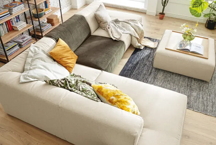 Helles Ecksofa mit grünem Sitzelement und gemusterten Kissen, davor quadratischer Polsterhocker als Couchtisch mit Tablett und Vase, hinten Regal mit Büchern und Pflanzen.
